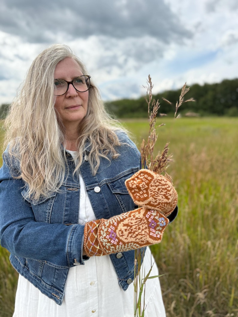 May include: A person wearing a denim jacket and holding a pair of knitted mittens. The mittens feature a brown, tan, and purple floral design. The background is a field of tall grass under a cloudy sky.