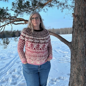 May include: A woman wearing a pink and red patterned jumper with a white background and blue jeans. She is standing in a snowy field with a tree behind her.