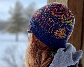 May include: A person wearing a blue knitted hat with a colorful geometric pattern. The hat has a red, yellow, and orange design. The person is wearing a gray hooded sweatshirt and looking out at a snowy landscape.