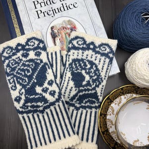 Puede incluir: Un par de guantes sin dedos de punto marfil y azul marino con un patrón detallado, junto a un ejemplar de "Orgullo y Prejuicio", una madeja de lana azul marino y una taza de té sobre un plato decorativo. Los guantes tienen un borde festoneado.