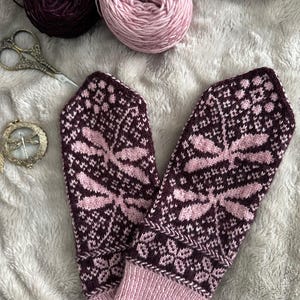 May include: A pair of knitted fingerless gloves with a dragonfly pattern in pink and burgundy. The gloves are made with a fine yarn and have a delicate, intricate design.