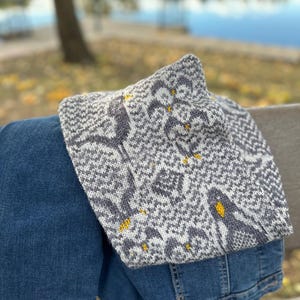 Può includere: Una sciarpa di lana grigia e bianca con un motivo geometrico e accenti gialli. La sciarpa è drappeggiata su un paio di jeans blu.
