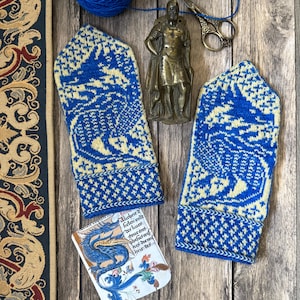 Puede incluir: Un par de mitones de punto azules y crema con un patrón detallado. El diseño incluye un motivo de dragón. La imagen también muestra hilo, tijeras, una pequeña estatua y una tarjeta decorativa con una ilustración de dragón y texto.