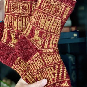 Könnte beinhalten: Ein Paar wadenhohe Strickstrümpfe in den Farben Dunkelrot und Goldgelb. Die Socken zeigen ein komplexes geometrisches Muster mit sich wiederholenden rechteckigen Motiven und stilisierten Tierfiguren, die in das Strickdesign integriert sind.