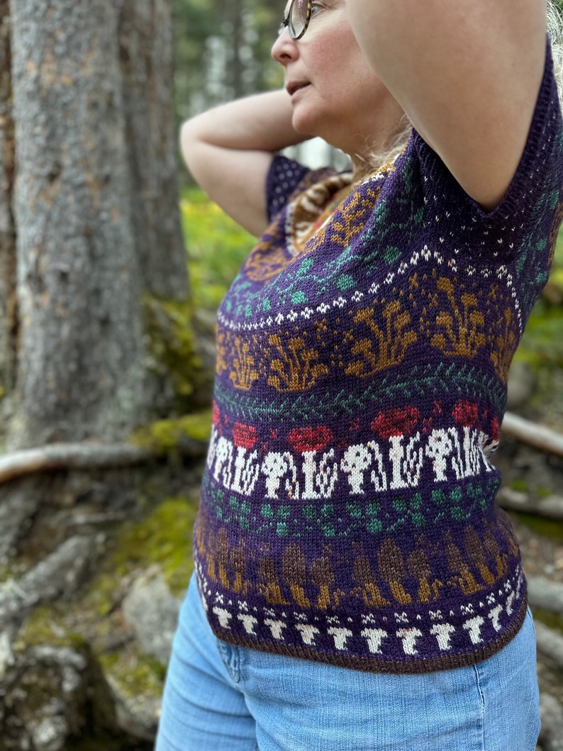 Pode incluir: Um su&eacute;ter de malha roxo, marrom e verde com um design de cogumelos e floresta. O su&eacute;ter tem mangas curtas e um decote em V.