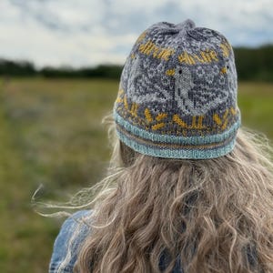 Puede incluir: Un gorro de punto gris con un dise&ntilde;o amarillo y blanco. El gorro tiene un borde azul claro y marr&oacute;n. El gorro lo lleva una persona con pelo largo y rubio y una chaqueta vaquera. El fondo es un campo y un cielo nublado.