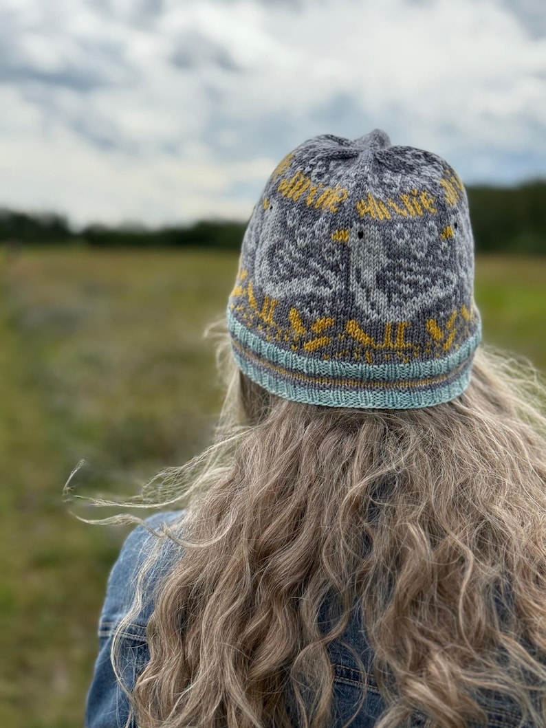 Puede incluir: Un gorro de punto gris con un dise&ntilde;o amarillo y blanco. El gorro tiene un borde azul claro y marr&oacute;n. El gorro lo lleva una persona con pelo largo y rubio y una chaqueta vaquera. El fondo es un campo y un cielo nublado.