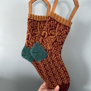 Puede incluir: Un par de calcetines de punto con un patrón marrón, naranja y turquesa. Los calcetines están colgados en perchas de madera.