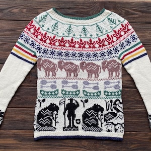 May include: Cream-colored knitted sweater with a colorful geometric pattern. The design includes red maple leaves, blue snowflakes, brown elephants, and black and white figures. The sleeves have striped accents and floral cuffs.
