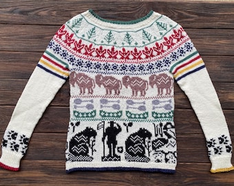 May include: Cream-colored knitted sweater with a colorful geometric pattern. The design includes red maple leaves, blue snowflakes, brown elephants, and black and white figures. The sleeves have striped accents and floral cuffs.