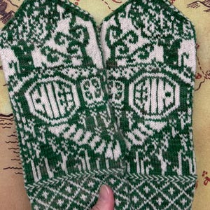 May include: A pair of green and white knitted mittens with a detailed pattern. The mittens feature a geometric design at the cuff and a central emblem. The overall design is intricate, with a combination of solid and patterned areas. The mittens are held by a hand.