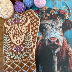 Könnte beinhalten: Ein Paar braune und beige gestrickte Fäustlinge mit floralen und geometrischen Mustern. Bunte Wollknäuel in Braun-, Beige-, Lila-, Blau- und Rosatönen sind um die Fäustlinge angeordnet. Eine Kuh-Illustration mit Blumenkranz ist im Hintergrund.