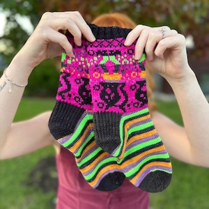 Puede incluir: Un par de calcetines tejidos a mano con una vibrante paleta de colores en rosa neón, negro, verde brillante y naranja. La sección superior muestra un patrón detallado de siluetas negras sobre fondo rosa neón, mientras que la inferior presenta rayas horizontales en negro, verde y naranja.