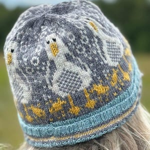 Puede incluir: Un gorro de punto gris con un diseño de gansos blancos con picos y patas amarillas. El gorro tiene un borde a rayas azules y amarillas. Los gansos están dispuestos alrededor del gorro. El gorro está hecho de un material de punto suave.