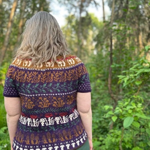 Pode incluir: Um su&eacute;ter de manga curta roxo, verde, marrom e branco com um design tem&aacute;tico de floresta que apresenta cogumelos, folhas e outros elementos da floresta.
