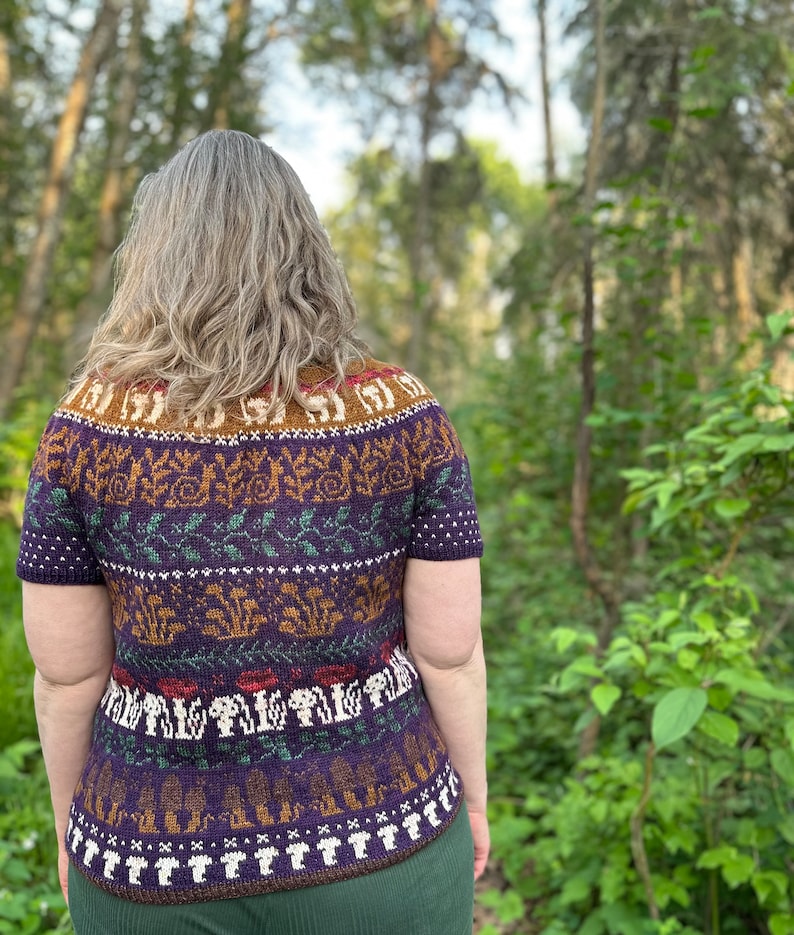 Pode incluir: Um su&eacute;ter de manga curta roxo, verde, marrom e branco com um design tem&aacute;tico de floresta que apresenta cogumelos, folhas e outros elementos da floresta.