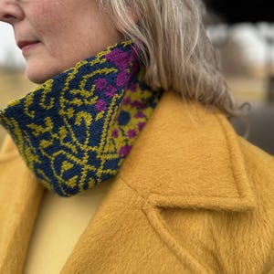 May include: A woman wearing a yellow coat with a blue, green and purple patterned knitted neck warmer.