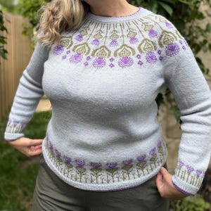 以下が含まれることがあります： 紫とオリーブグリーンの花柄が特徴の、水色のセーター。セーターは丸いネックラインと、お揃いの袖口が付いた長袖が特徴です。花柄は裾とネックラインの周りに繰り返されています。