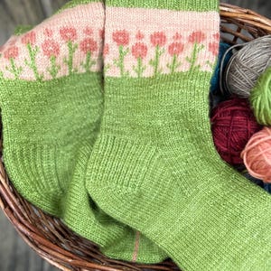 May include: A pair of green knit socks with a floral pattern. The top of the socks features a pink band with pink flowers and green stems. The socks are in a wicker basket with balls of yarn.