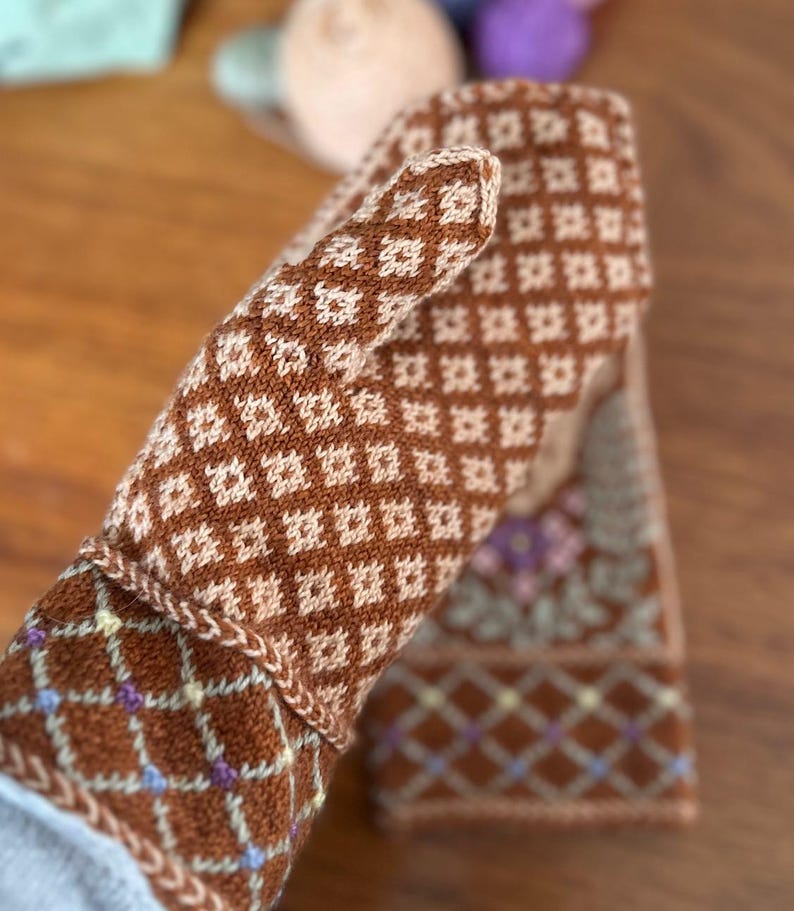 May include: A pair of brown and cream-colored knitted mittens with a diamond pattern. The mittens have a floral design on the cuff and are made of a soft, textured yarn. The cuff features a diamond pattern with small accents of purple and yellow.