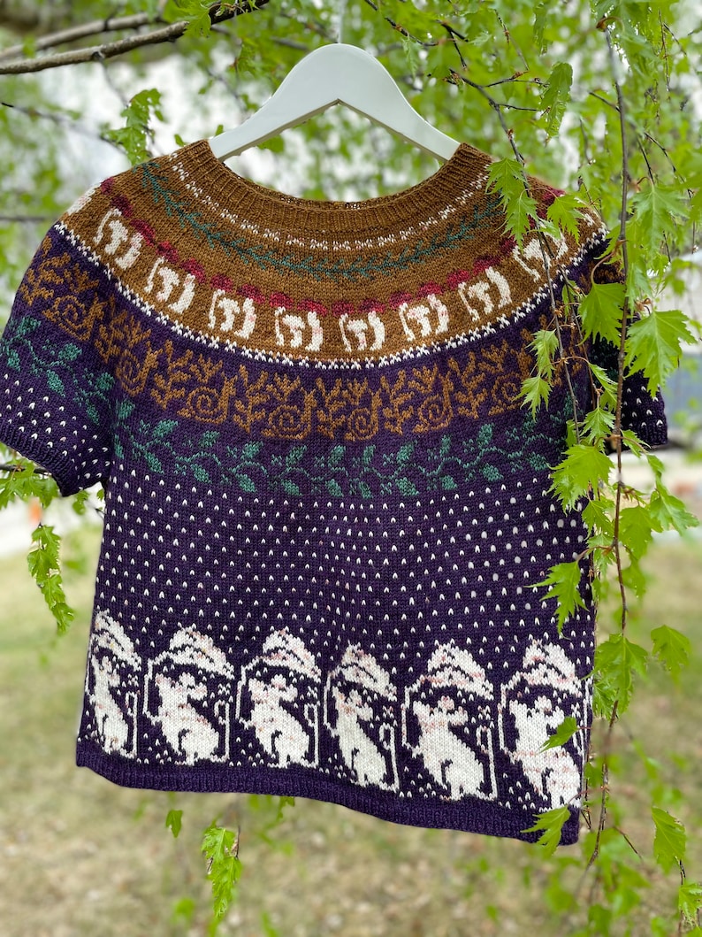 Pode incluir: Um su&eacute;ter de malha roxo, verde, marrom e branco com um design de manga curta. O su&eacute;ter apresenta um padr&atilde;o repetitivo de animais brancos em uma floresta nevada.