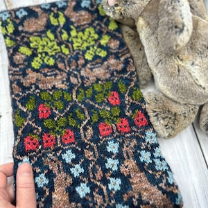 Può includere: Una sciarpa di maglia con un motivo dettagliato di alberi, foglie e bacche in tonalità di verde, rosso, marrone e blu.