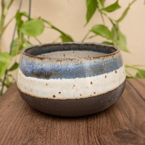 May include: A handmade ceramic bowl with a dark brown base, a cream-colored middle section, and a blue top. The bowl is round and sits on a wooden surface. The bowl is approximately 4 inches in diameter.