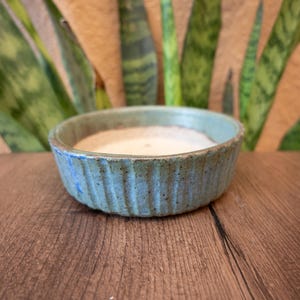 May include: A round, shallow ceramic bowl with a textured, ribbed exterior in shades of muted blue and green. The interior of the bowl is a smooth, off-white color. The vessel has a rustic, speckled finish across its outer surface.