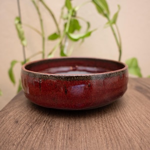 May include: A handmade, round ceramic bowl with a glossy, deep red glaze. The bowl has a dark brown rim and sits on a wooden surface. The bowl is ideal for serving food or as a decorative piece.