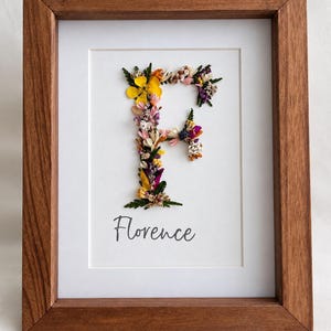Könnte beinhalten: Ein rechteckiger Holzrahmen mit einem weißen Passepartout, das ein aus gepressten Blumen geformtes F enthält. Der Buchstabe besteht aus verschiedenen kleinen, bunten Trockenblumen und grünen Blättern. Darunter ist der Name Florence aufgedruckt.