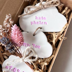 May include: Three white cloud-shaped ornaments with pink embossed text including the name Kate Phelan and a date. The ornaments are tied with brown twine and decorated with a small bouquet of dried pink and purple flowers inside a cardboard box.