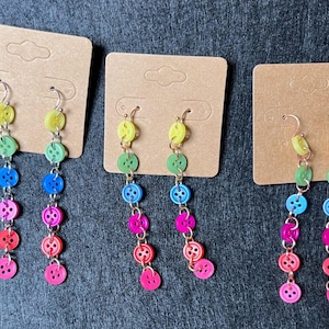 May include: Three pairs of dangle earrings featuring a rainbow of coloured buttons. Each earring has a silver hook and a chain with five buttons in different colours: yellow, green, blue, pink and red.