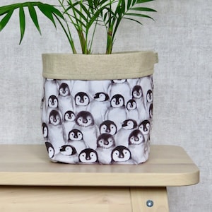 May include: A white fabric planter with a beige linen top rim. The planter is decorated with a pattern of black and white penguins. A green potted plant is inside the planter.