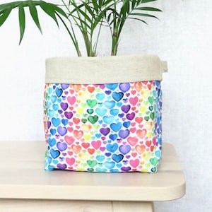 May include: A white fabric planter with a rainbow heart pattern. The planter has a beige linen top and is filled with a green plant.