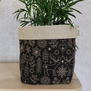 May include: A black fabric plant pot with a beige rim, decorated with a festive pattern of snowflakes, trees, and reindeer. The pot contains a green leafy plant, ideal for home decor.