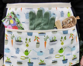 May include: A white gardening apron with a colorful pattern of plants, watering cans, and carrots. The apron's pockets hold green gardening gloves, seed packets, wooden plant markers, and twine. The apron is designed for gardening.