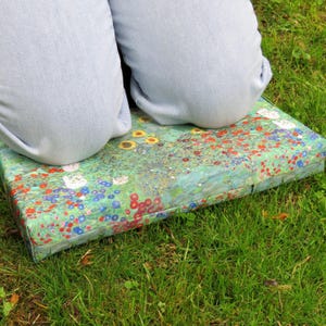 May include: A rectangular cushion with a floral design featuring red, yellow, and blue flowers on a green background. The cushion is placed on green grass, with two light blue denim-clad legs resting on top.
