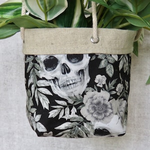 May include: A black and white fabric hanging planter with a skull and floral design. The planter is made of linen and has a drawstring closure.