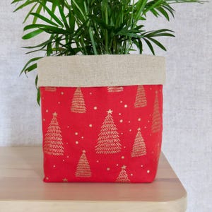 May include: A red fabric plant pot with a gold Christmas tree pattern. The top edge is a natural linen colour. The pot holds a green plant with long, thin leaves. The pot sits on a light-coloured wooden surface.