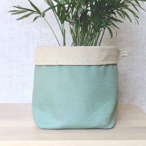 May include: A light green canvas planter with a cream coloured top rim. The planter is filled with a green leafy plant.
