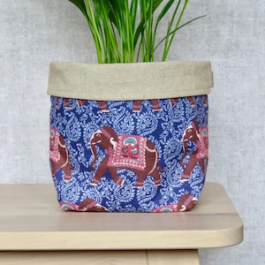 Peut inclure: Un pot de fleurs en tissu bleu avec un dessus en lin beige. Le pot présente un motif répété d'éléphants bruns avec des accents roses et rouges sur un fond bleu paisley.