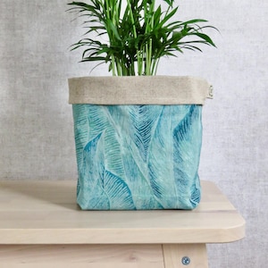 Peut inclure: Un pot de plante carré en tissu avec un motif de feuilles turquoise. Le bord supérieur est de couleur beige neutre. Le pot contient une plante verte avec de longues feuilles fines. Le pot est posé sur une surface en bois de couleur claire.