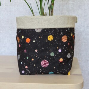 May include: A black fabric planter with a beige linen top. The planter features a repeating pattern of planets and stars on a black background.