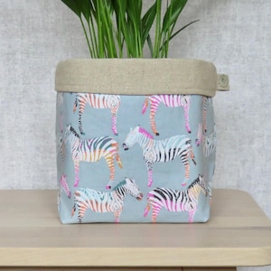 May include: A light blue fabric storage basket with a beige linen top. The basket features a colourful zebra print pattern.