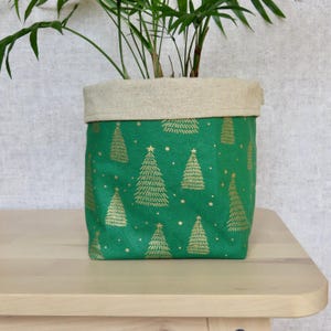 May include: A green fabric plant pot featuring a gold Christmas tree design. The top edge is a natural beige colour. A green plant is inside the pot, on a light wood surface.