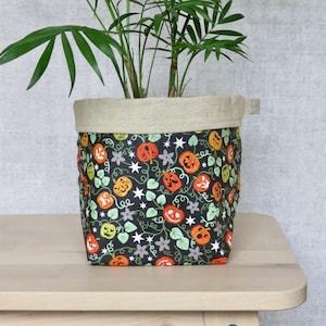 May include: A fabric plant pot cover featuring a black background with a repeating pattern of orange jack-o'-lanterns, green leaves, and white stars. The top edge of the container is made of a plain, light beige fabric that folds over the rim.