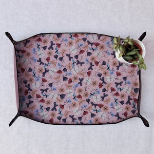 May include: A pink tray with a black border and a pattern of butterflies and flowers. The tray is made of fabric and has a slightly rounded shape.