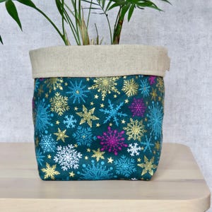 May include: A teal blue fabric planter with a gold and white snowflake pattern. The planter has a natural linen top and is filled with a green plant.