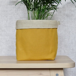 May include: A mustard yellow fabric planter with a natural linen top. The planter is sitting on a light wood surface and has a plant growing inside.