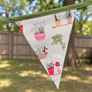 Peut inclure: Un fanion triangulaire en tissu avec un fond gris clair et un motif répétitif de plantes et d'herbes en pot. L'imprimé présente des illustrations de ciboulette, de fraises, de romarin et de lierre dans des pots colorés avec des étiquettes.